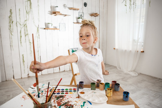 Adorable Caucasian little creative and talanted girl holding big brush in her hand and deeping it into water, standing behind desk in the art room. Cute preschool blonde female child wearing white