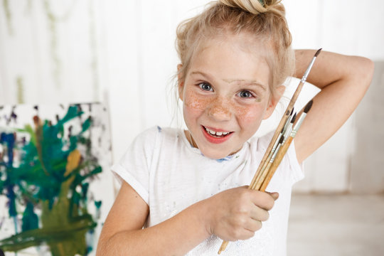 Portrait of angel-like cheerful smiling with teeth child in white morning light in art room, holding in her hand bunch of brushes. Little European girl with blond hair looking happy and joyful showing