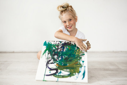 Close-up portrait of blond European little girl with hair bun and freckles smiling with all her teeth. Holding on her knees picture that she painted for her parents, feeling proud of herself. People