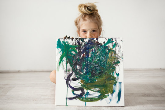 Studio shot of beautiful little blonde girl holding and hiding by the big picture, looking at the camera with cunny and joyful expression. Cute child with hair bun, freckles and blue eyes showing