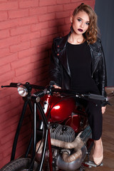 Fototapeta na wymiar Coveted woman or girl in a leather jacket and tight pants, boots sits on a motorcycle, with an unusual hairdo and make-up on a brick red wall