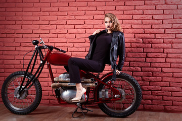 Obraz na płótnie Canvas Coveted woman or girl in a leather jacket and tight pants, boots sits on a motorcycle, with an unusual hairdo and make-up on a brick red wall