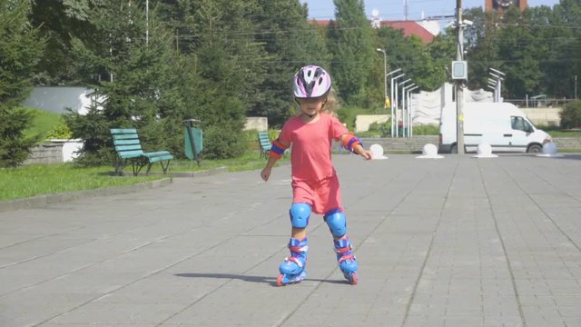 A little girl in a helmet and defense skates on roller skates. The child rolls on the rollers in the park.  Rollerblading In The Park.  Girl learns to ride a roller skate.