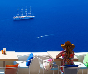Woman in hat sitting in cafe and enjoys views of sailing cruise ship, Santorini, Fira, Greece