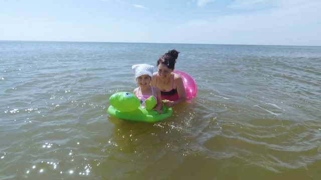  The mother and her little daughter bathe and play in the sea in the water. Mother with  kid swim in wavy sea, holding green inflatable ring sunny day.