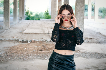 A coveted woman or girl stands in the middle of an abandoned building in a leather skirt top and sunglasses, sexy brunette