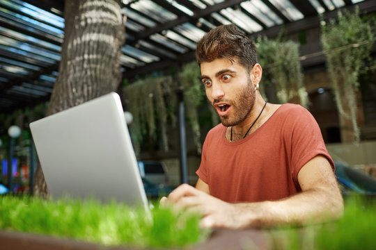 Shot of shocked bearded young man with dark hair and popped eyes, opened mouth dressed in red t-shirt looking at the screen of his notebook recieving e-mail with important news.