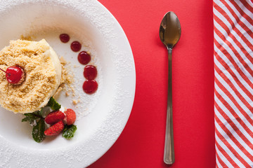 Napoleon cake serving on red background. Sweet dessert on plate with decoration from strawberry, cherry and mint, close up top view, free space on right