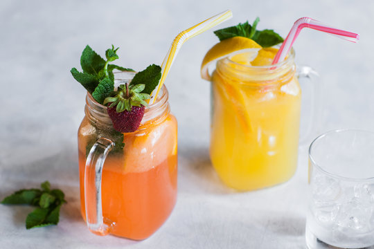 Fresh fruit cocktails in jars on white table. Orange and strawberry cold drink with mint and glass with ice close by, concept of refreshment in hot weather
