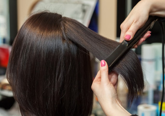 The hairdresser does aligns the hair with hair iron to a young girl, brunette in a beauty salon. Professional Hair Care