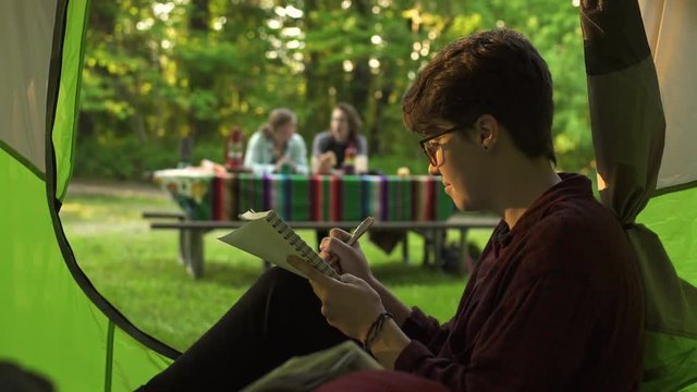 Camper Sits In Tent And Writes In Journal, Friends Sit At Picnic Table In Background