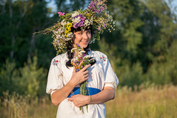 A young woman in a long white embroidered shirt and in a wreath of wild flowers holds a bouquet of flowers in his hands in a meadow at sunset.
