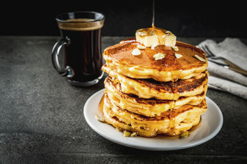 Autumnal traditional food. Stack of pumpkin pancakes with butter, pumpkin seeds and maple syrup. With a cup of coffee. On a black stone table. Copy space