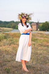 Fototapeta na wymiar A young woman in a long white embroidered shirt and in a wreath of wild flowers stands in the field at sunset.