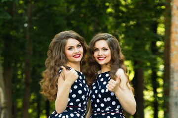 Two beautiful smiling girl sisters twins for a walk in summer park on a background of greenery with makeup and hairstyle