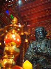 Close up old black monk man statue, respected in Chinese wooden red shrine, with blurred lamp foreground