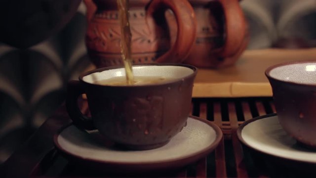 The action of traditional Chinese tea ceremony. Pouring tea from clay tea pot in a cup. Two tea pots in soft focus on the background.