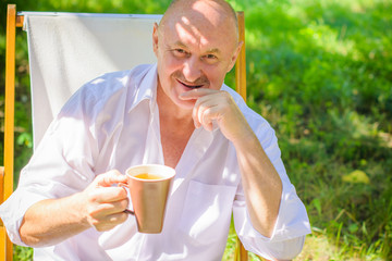 Mature European or American man in white shirt with cup of tea sits in armchair and rest in garden at morning feeling happy and comfortable. 
