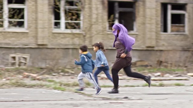 Tracking of laughing middle-eastern children and woman in purple burqa running through street with abandoned building