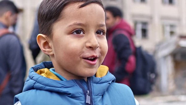 PAN of cheerful Arab refugee boy with dirt on his face wearing puffy vest and talking, then looking at camera and smiling; people with backpacks standing in background