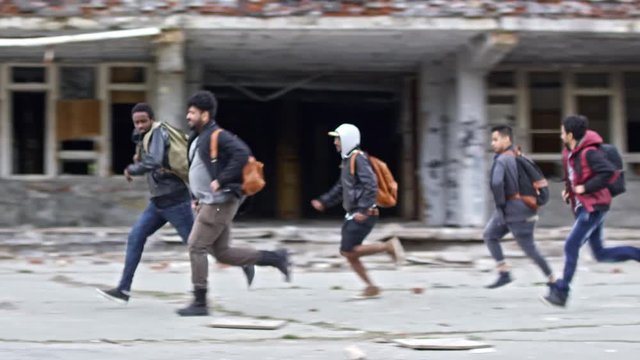 Tracking of group of Arab refugees running before abandoned building with broken windows