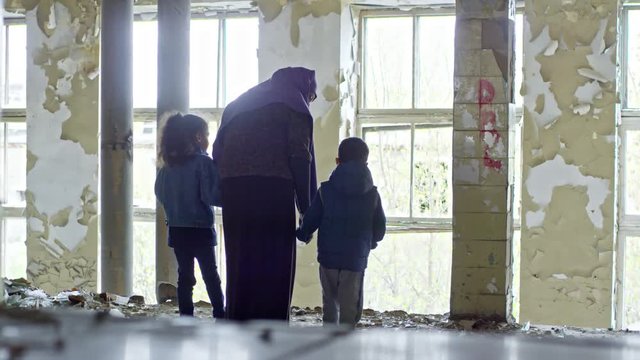 PAN with rear view of Muslim woman in purple niqab holding hands of her little children and looking out window of abandoned house