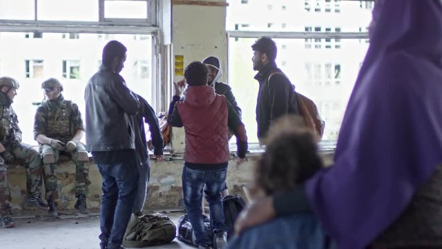 PAN of unrecognizable Muslim woman in purple niqab and little child looking at happy Arab male refugees dancing in destroyed building; Caucasian soldiers in uniform chatting in background