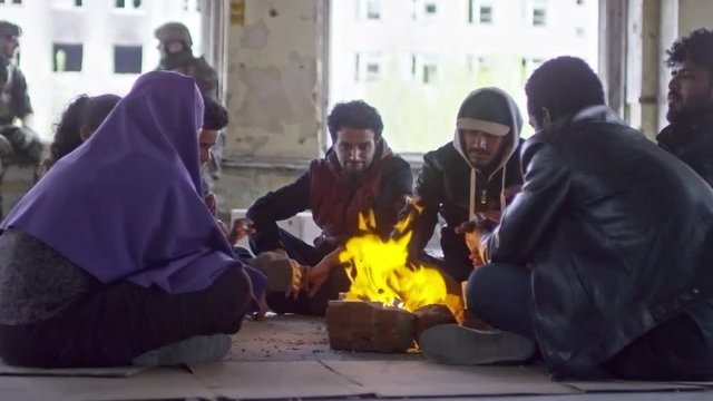 PAN of female Muslim refugee in purple niqab, little boy and group of Arab men sitting before fire in abandoned building;  soldiers sitting on windowsill of broken window and chatting in background
