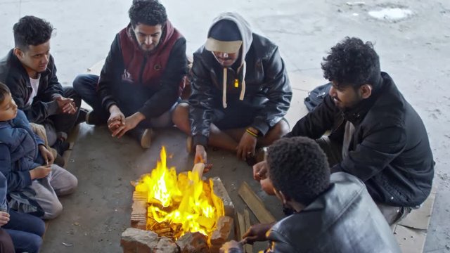 PAN with top view of group of Arab men in jackets and Muslim woman in niqab with little girl sitting in circle before fire and chatting