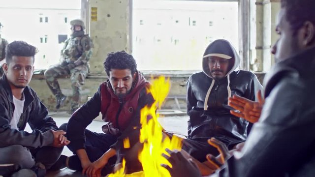 PAN of Arab men in jackets, Muslim woman in purple niqab and little children sitting by fire in abandoned building; soldiers in uniforms sitting on windowsill of broken window and chatting
