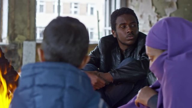 PAN of Muslim woman in niqab and little boy sitting by fire in abandoned building and chatting
