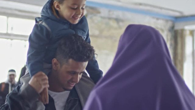 Tilt down of little boy sitting on shoulders of cheerful young Arab man chatting with unrecognizable woman in purple niqab