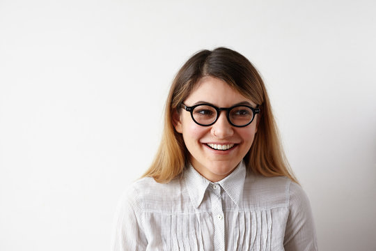 Headshot of beautiful positive young Caucasian woman wearing stylish eyeglasses and blouse standing at white office wall and smiling broadly during job interview with human recources specialist