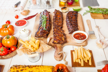 Spicy hot grilled spare ribs from a summer BBQ served with chips, corn and fresh tomatoes on an old wooden cutting board