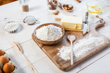 Baking ingredients in rural kitchen - dough recipe ingredients (eggs, flour, milk, butter, sugar) and rolling pin on wooden white table.