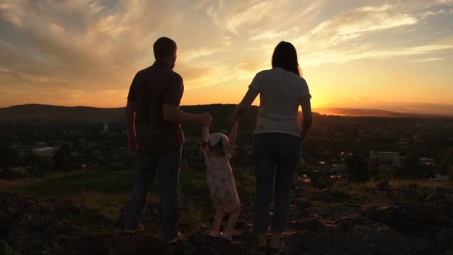 The family is standing on a hill at sunset on a summer evening, mom and dad are holding a small daughter's hands. A little girl is jumping between her parents.