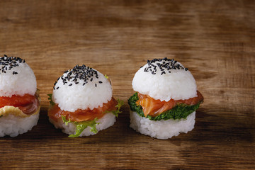 Mini rice sushi burgers with smoked salmon, green salad and sauces, black sesame served with wooden chopsticks over wood background. Modern healthy food