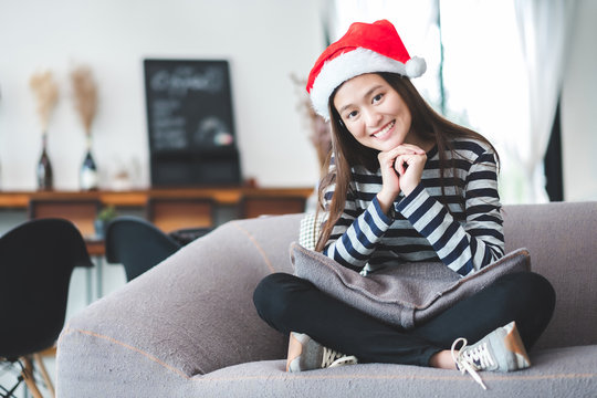 Asian teenage girl sitting cross-legged on sofa with Christmas hat and smile face and  resting her chin on her hands at home ,Celebrate New year and Christmas holiday concept