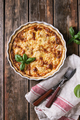 Baked homemade quiche pie in white ceramic form served with fresh greens, cutlery and textile towel on old wooden plank background. Flat lay with copy space