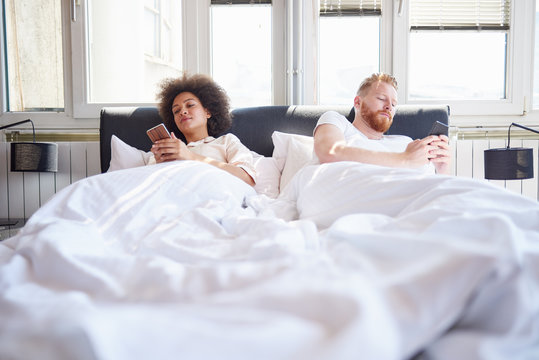 Young couple using mobile phones and lying in bed while ignoring each other