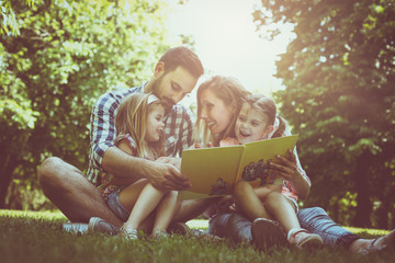 Happy family with two children in meadow reading book together.