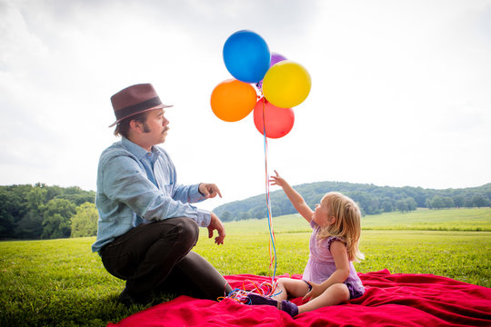 Girl sitting with father pointing up to bunch of colourful balloons in rural field