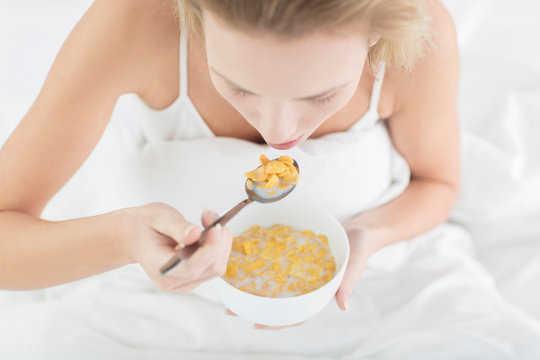 Young woman sitting in bed, eating bowl of cereal, elevated view