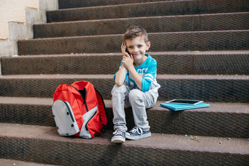 Smiling child making a phone call outside a school. Happy little student sharing great news over a...