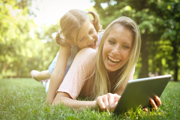 Mother and daughter outdoors in a meadow. Mother with her daughter lying on grass and using digital tablet together.
