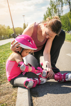 Smiling mother with her little daughter sitting on road after skating. Mother comforting daughter