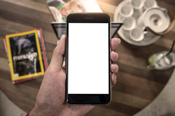 Close up of a man holding smartphone with blank screen mobile and laptop computer at coffee shop.
