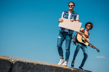 multicultural men with empty cardboard walking on parapet while hitchhiking together