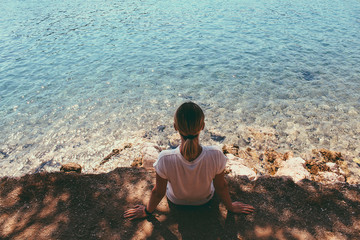 Back view of sitting traveler, holding the arms on the shore, enjoying the landscape of sparkling sea water and rocks.