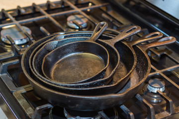 An iron skillet collection, ready to start cooking, stacked on top of a stovetop with a selective focus on the edge of the pans.